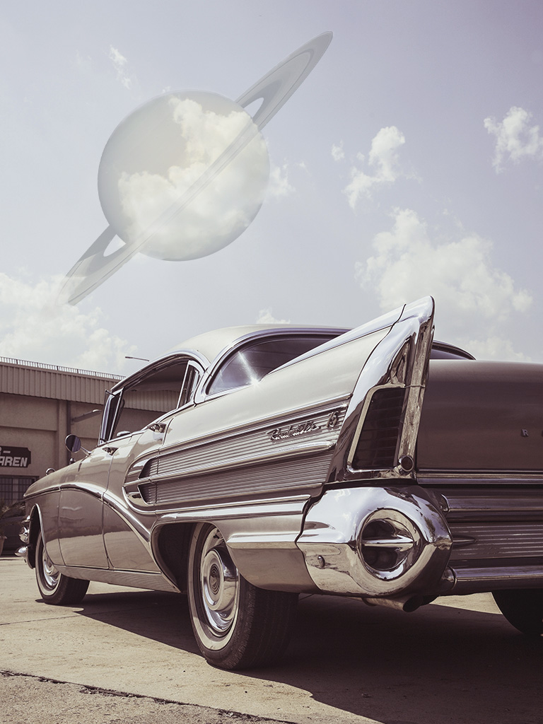 A classic 1950s Chevy Bel Air dominates the foreground, facing away so that the fins are prominent. In the sky above, a ghostly, ringed planet is partially hidden by clouds. Creative Commons images Photoshopped by Jonny Eberle.
